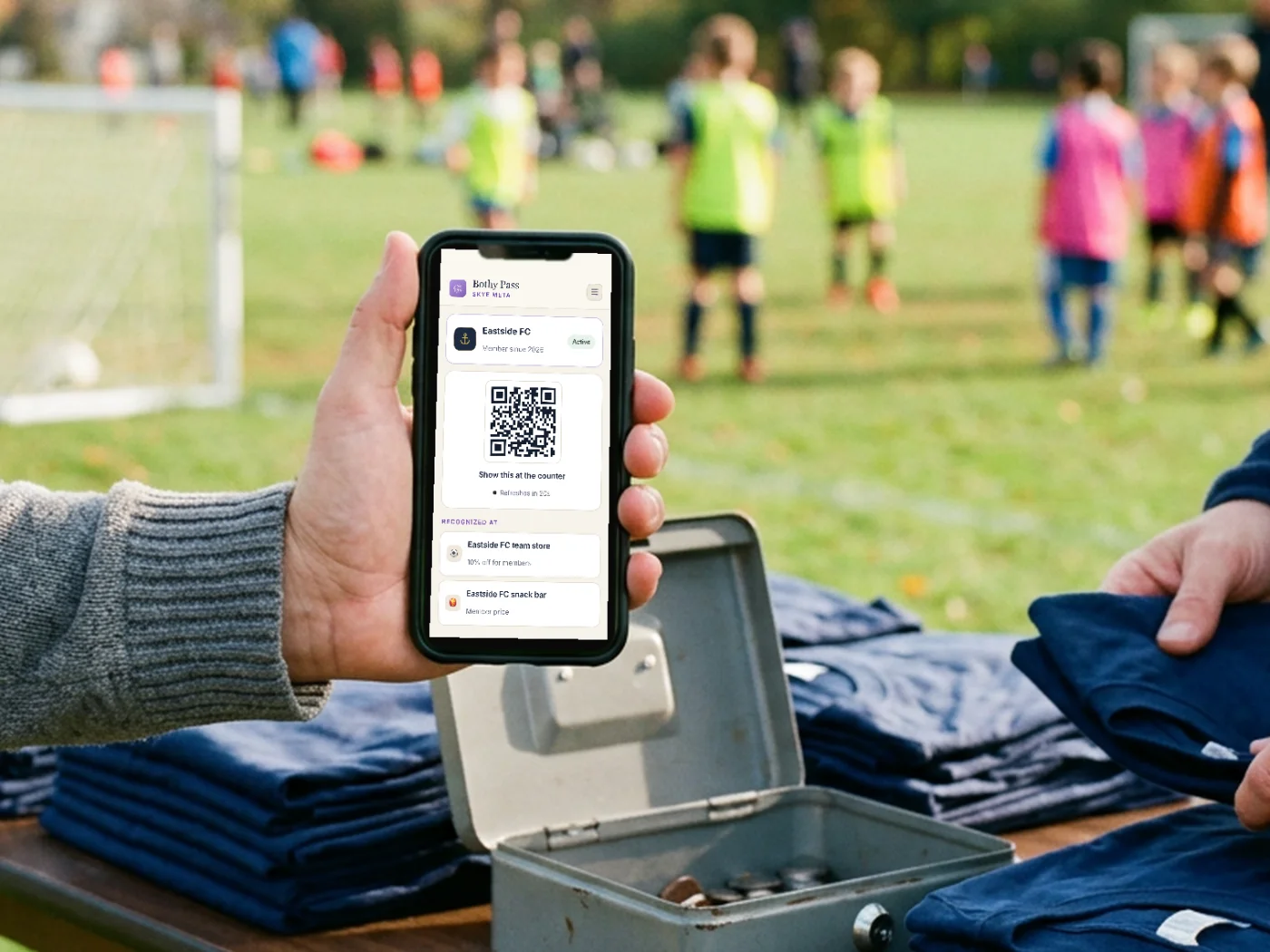 A parent holds up a Bothy Pass on their phone at a team-store table beside a youth soccer pitch, with folded club t-shirts and a cash box on the table